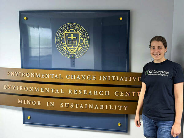 Photo of Lisa Schulz standing in front of a sign for the Environmental Change Initiative | Environmental Research Center | Minor in Sustainability. Lisa's shirt reads "CI Compass Fellowship Program."