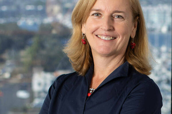 A woman with shoulder-length blond hair smiles at the camera with her arms crossed. She wears a dark blue, button-down shirt, a fitness tracker, and red earrings and necklace. A blurred cityscape is visible through the window behind her.