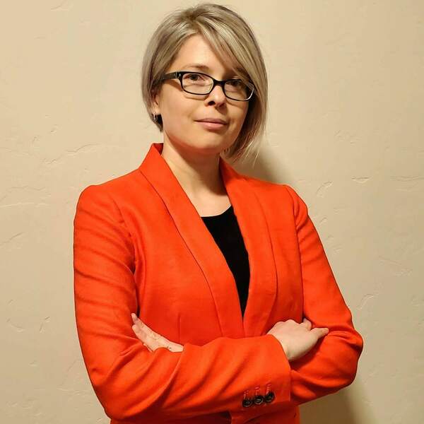 Headshot of Svitlana Volkova, who has blonde hair, wears glasses, and is wearing a deep orange jacket.