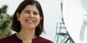Woman with short brown hair, wearing a maroon shirt, smiles at the camera with a large satellite dish in the background.