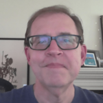 Headshot of a man with glasses and a blue shirt, slightly smiling. Artwork and framed photos are visible on the wall behind him.