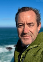 Man in a green fleece jacket smiles at the camera with a blue ocean and white waves in the background.
