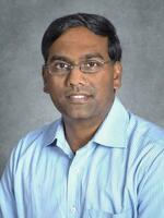Headshot of a person with dark hair, wearing glasses and a light blue pinstriped shirt, against a gray background.