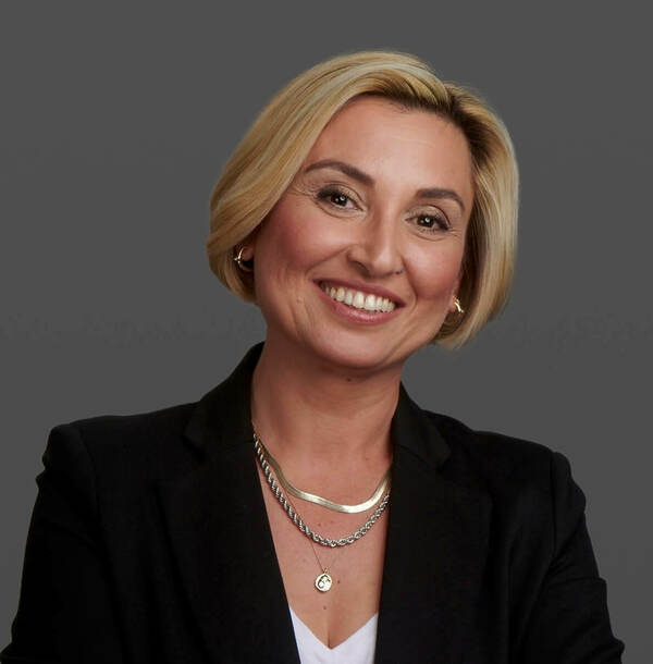 Woman with short blond hair, wearing a black blazer and silver necklace, smiles against a gray background.