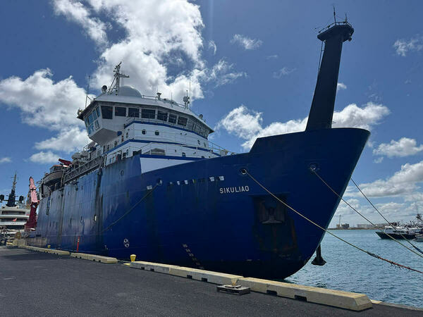 Dark blue research vessel Sikuliaq, with white superstructure and a tall black mast rising from its bow, is docked at a pier under a bright, partly cloudy blue sky.