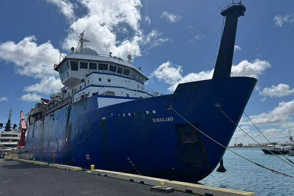 Dark blue research vessel Sikuliaq, with white superstructure and a tall black mast rising from its bow, is docked at a pier under a bright, partly cloudy blue sky.