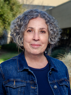 A woman with curly gray hair, wearing a denim jacket and hoop earrings, smiles gently outdoors against a blurred background of green foliage and a light building.