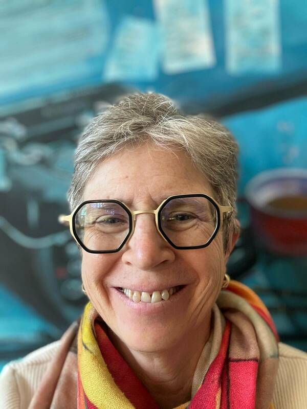 A smiling woman with short gray hair and octagonal black-and-gold glasses. She wears a cream sweater and a bright red, yellow, and orange scarf against a blurred blue background.