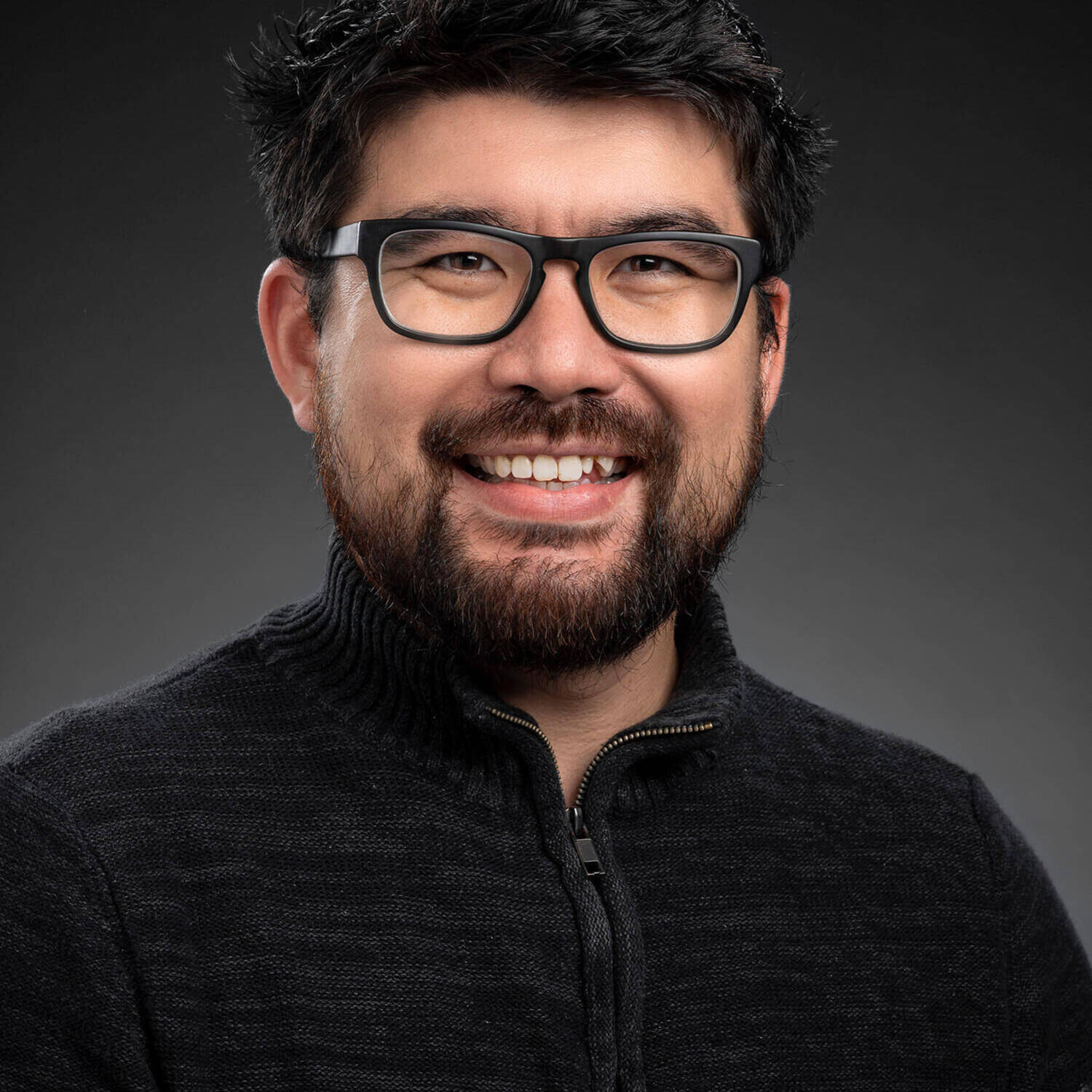 A man with dark hair, a beard, and black-rimmed glasses smiles broadly, wearing a dark gray quarter-zip sweater.