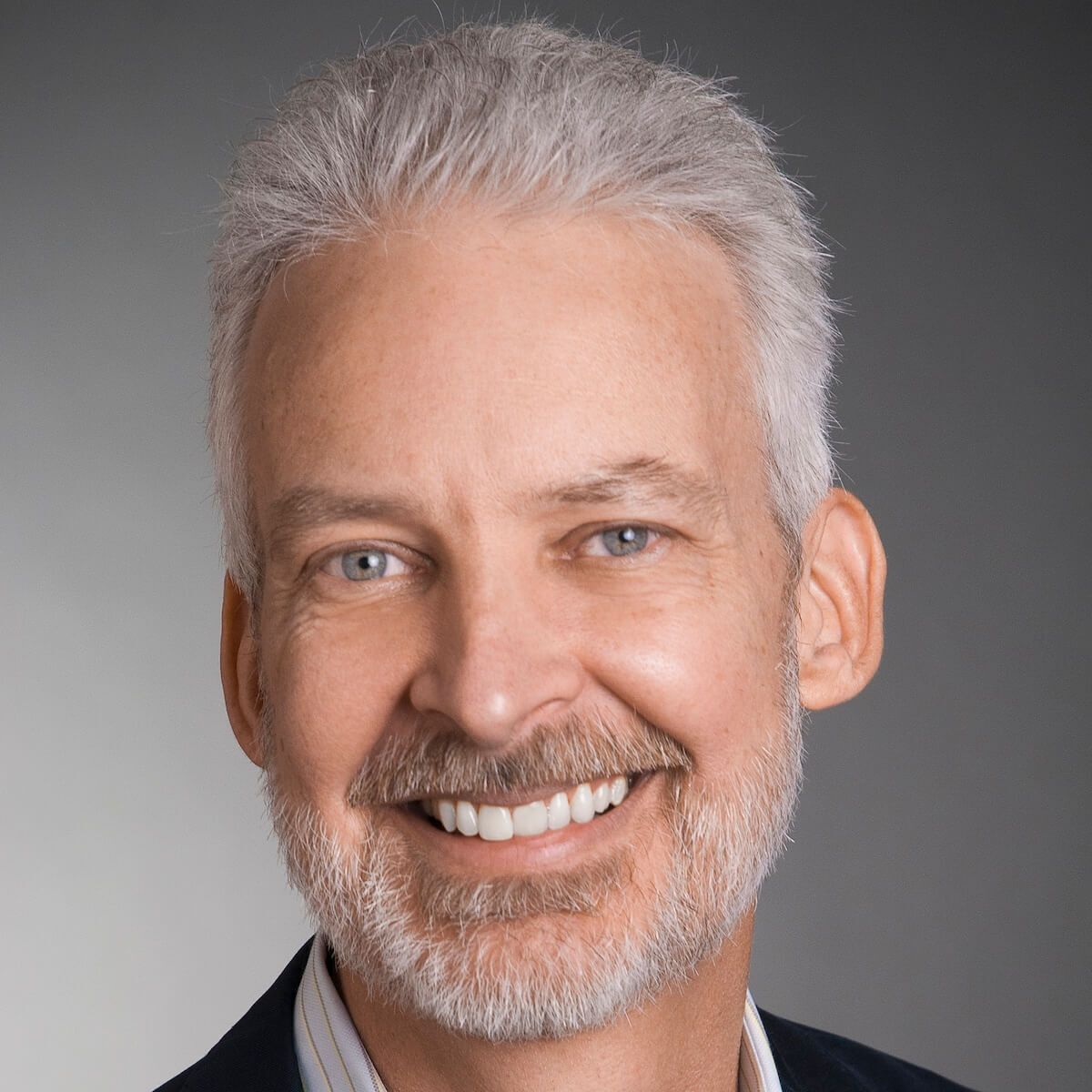 A smiling man with grey hair, a grey beard, and blue eyes wears a dark suit jacket and striped shirt against a grey background.