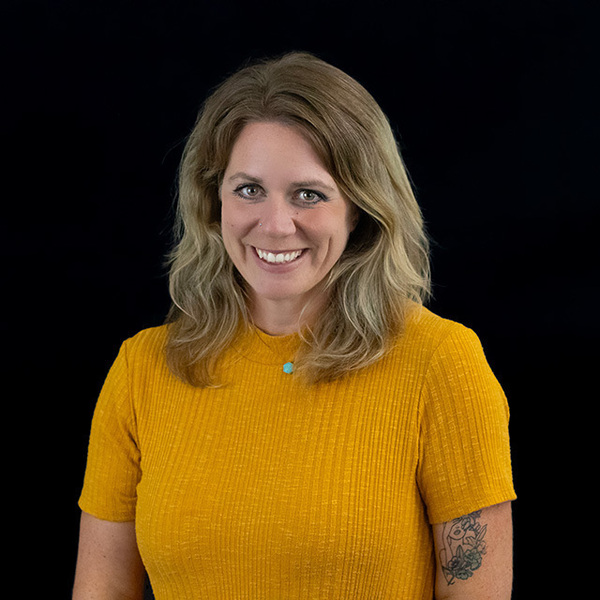 A woman with wavy blonde hair, green eyes, and a nose ring smiles warmly. She wears a yellow ribbed shirt and a small turquoise necklace against a black background.