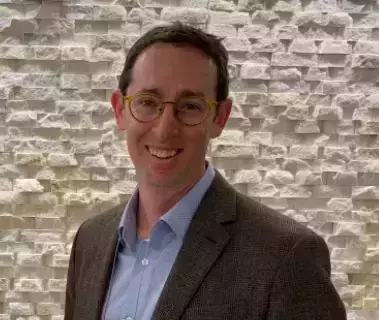 A man with short brown hair and round yellowish glasses smiles, wearing a light blue shirt and a brown plaid sport coat, against a light stone wall.