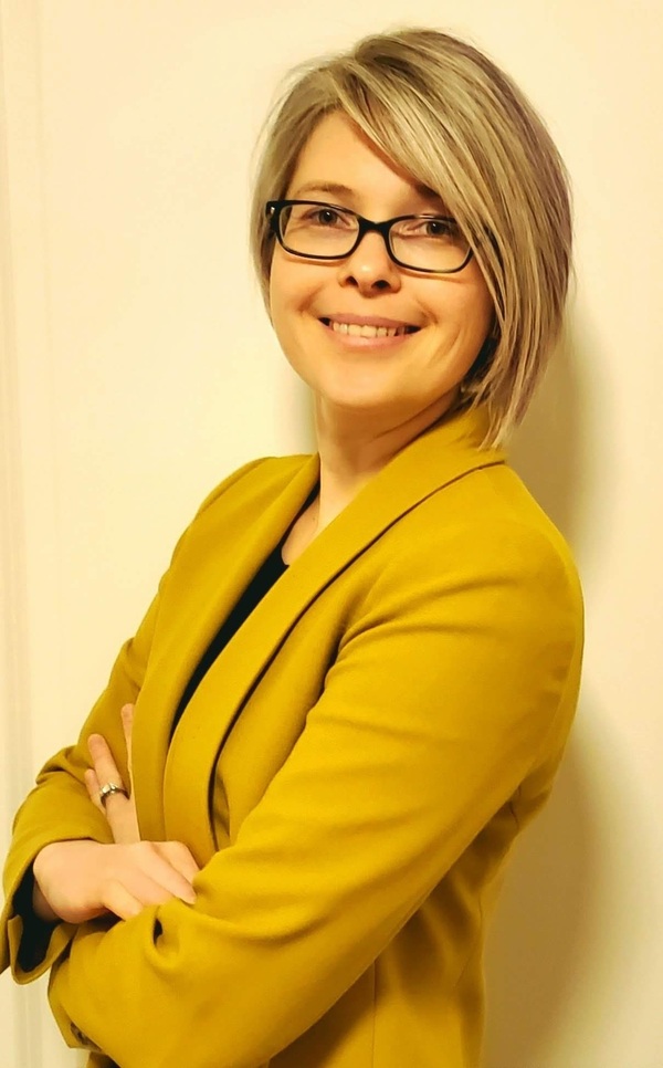 Smiling woman with short ash blonde hair and black-framed glasses, wearing a mustard yellow blazer. She stands with her arms crossed, looking directly at the viewer.