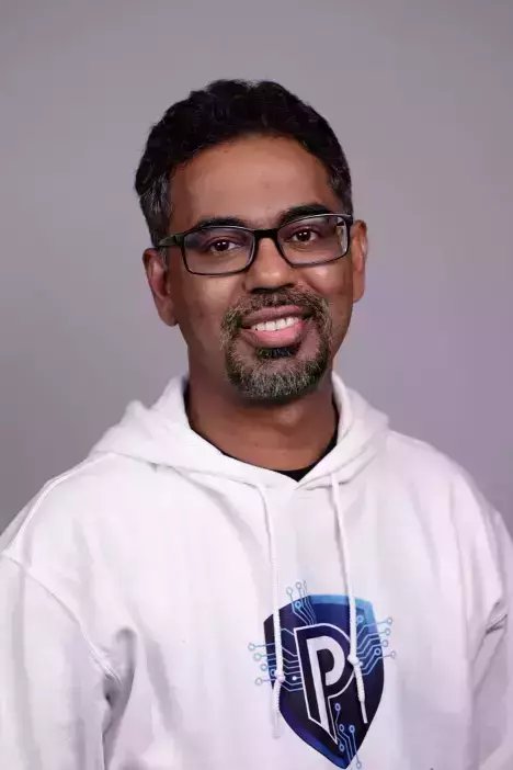 A smiling man with dark hair, a graying beard, and glasses wears a white hoodie with a blue and white shield logo.