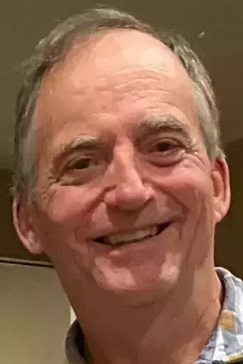Older man with graying brown hair and eyebrows smiling broadly, showing teeth. He wears a light blue and white plaid shirt.