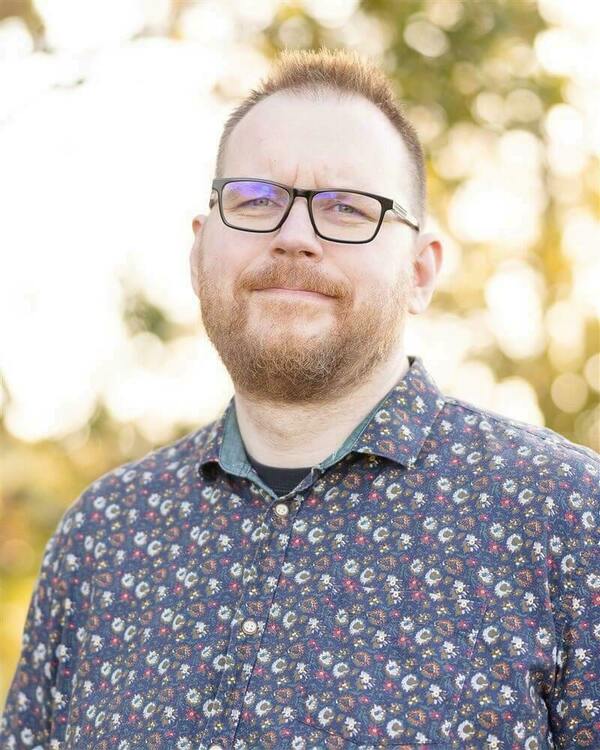 A man with a reddish-brown beard and black glasses smiles slightly, wearing a dark blue patterned shirt with small colorful floral designs. He stands outdoors with blurred green and yellow foliage behind him.