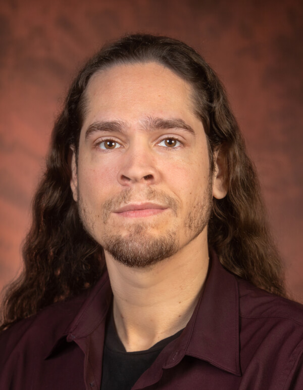 A man with long, wavy brown hair, a short beard, and a neutral expression looks forward. He wears a maroon collared shirt over a black t-shirt against a reddish-brown background.