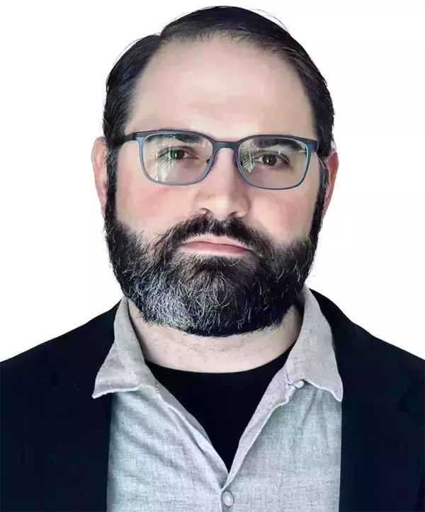 A man with dark hair, a dark beard with grey, and blue-rimmed glasses stares forward. He wears a black jacket over a grey collared shirt and black t-shirt.