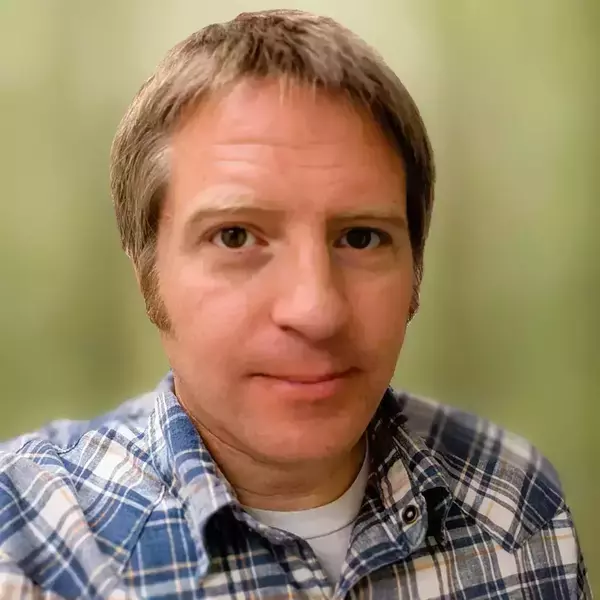 A man with short brown hair and brown eyes looks forward. He wears a blue, white, and tan plaid shirt against a light green blurred background.