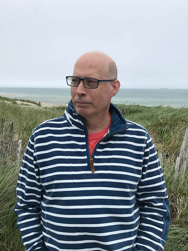 Bald man with blue glasses and a blue-white striped quarter-zip sweater over a red shirt stands, looking left, by a grassy dune. The ocean with a distant ship and an overcast sky are in the background.