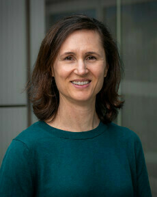 A smiling woman with shoulder-length brown hair wears a teal long-sleeved top, looking directly at the camera.