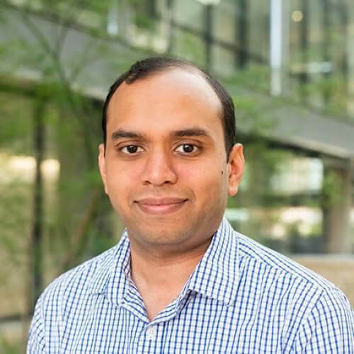 A man with light brown skin, dark hair, and a faint smile wears a blue and white checkered shirt against a blurry outdoor background of a modern building and green foliage.