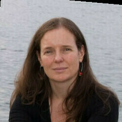 A woman with long wavy brown hair, wearing a black top and red dangle earrings, looks calmly at the viewer. A blurred grey body of water is behind her.
