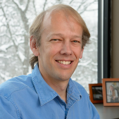 A fair-skinned man with light brown hair and a receding hairline smiles warmly in a light blue collared shirt. Behind him, a window reveals snow-covered trees. A blurred framed photo is visible on a desk to his right.