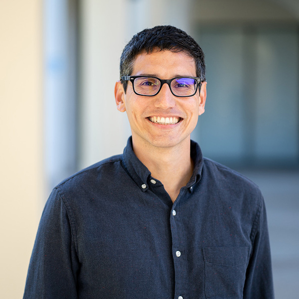 A man with short dark hair and black-rimmed glasses smiles broadly, showing teeth. He wears a dark blue button-up shirt against a blurred, light-colored architectural background.