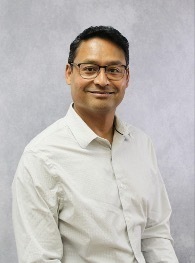 A man with dark hair and glasses smiles gently while wearing a light gray collared shirt against a plain gray background.