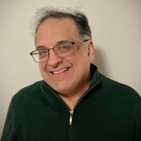 A man with graying dark hair and dark-framed glasses smiles broadly, showing teeth. He wears a dark green quarter-zip sweater.