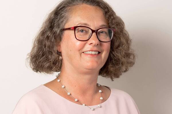 A smiling woman with wavy hair and red glasses, wearing a light pink top and a pearl necklace.