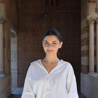 Young woman with dark hair, white shirt, and gold necklace smiles gently before a brick wall with ornate stone columns and arches.