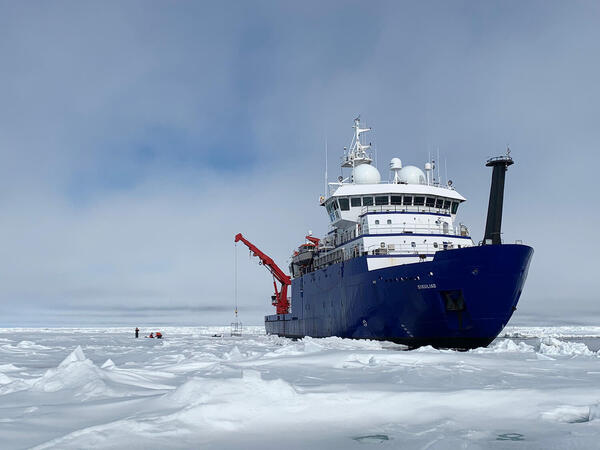 RV Sikuliaq, a research vessel that is dark blue on the bottom, white on top, and had red instruments off the stern, is seen in icy waters.
