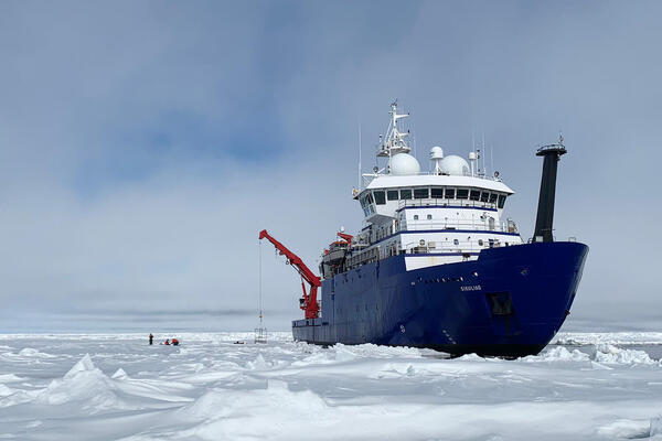 RV Sikuliaq, a research vessel that is dark blue on the bottom, white on top, and had red instruments off the stern, is seen in icy waters.