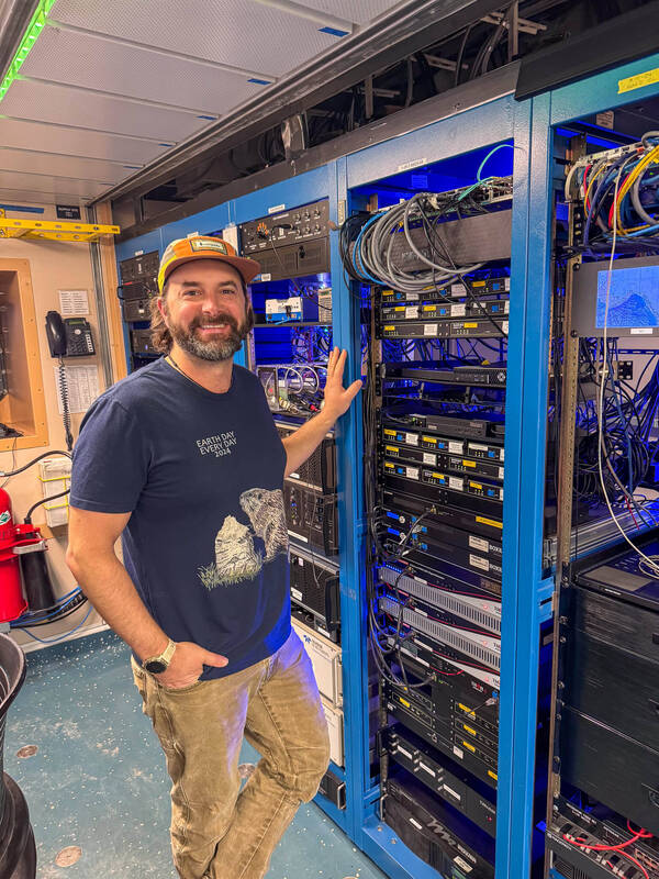 Julian Race stands in a navy blue t-shirt and tan pants next to a stack of computer servers in a lab on the ship.