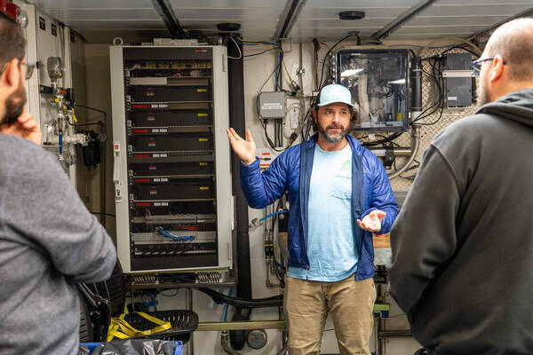 Julian Race, a bearded man in a blue jacket and light blue cap gestures, speaking to two men in front of a compact computer server on RV Sikuliaq.
