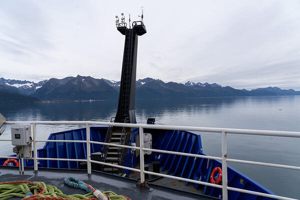 Ship's bow with blue hull, white railing, tall mast, facing calm blue water with distant snowy dark mountains and overcast sky.