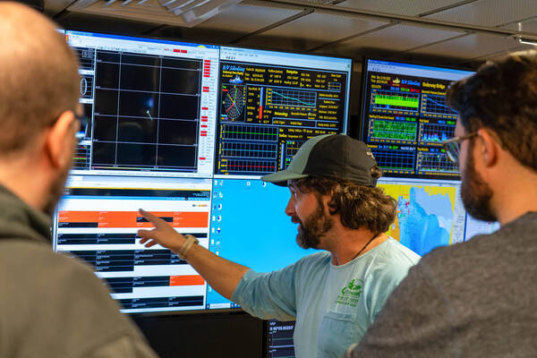 Julian Race, a man in a green cap, points at complex marine data on a ship's bridge monitor, with two colleagues observing screens.