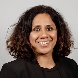 A smiling woman with dark, wavy hair and a black blazer against a light gray background.