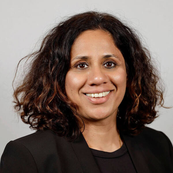 A smiling woman with dark, wavy hair and a black blazer against a light gray background.