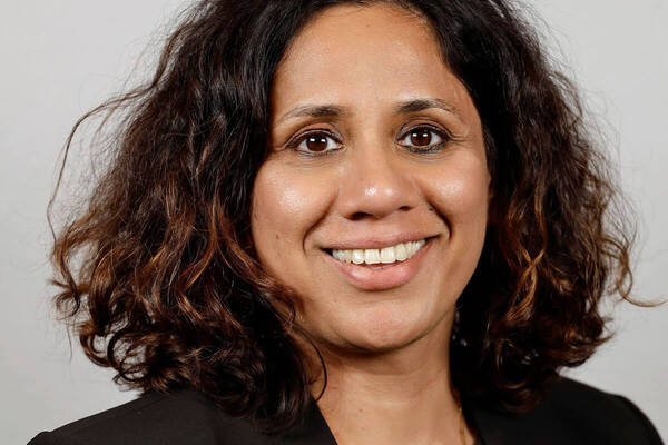 A smiling woman with dark, wavy hair and a black blazer against a light gray background.