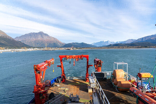 The research vessel deck of RV Sikuliaq, with large red cranes overlooks Resurrection Bay, in Seward, Alaska, with majestic mountains under a blue sky.