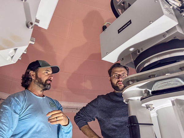 Two bearded men, one in a cap, intently observe large white telescope equipment in an observatory dome on RV Sikuliaq.