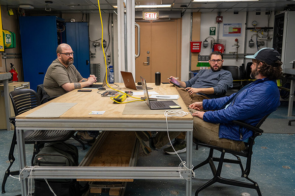 Three bearded men work on laptops at a large wooden table in a workshop in the lab on RV Sikuliaq. One man in blue speaks to another in a grey sweater.
