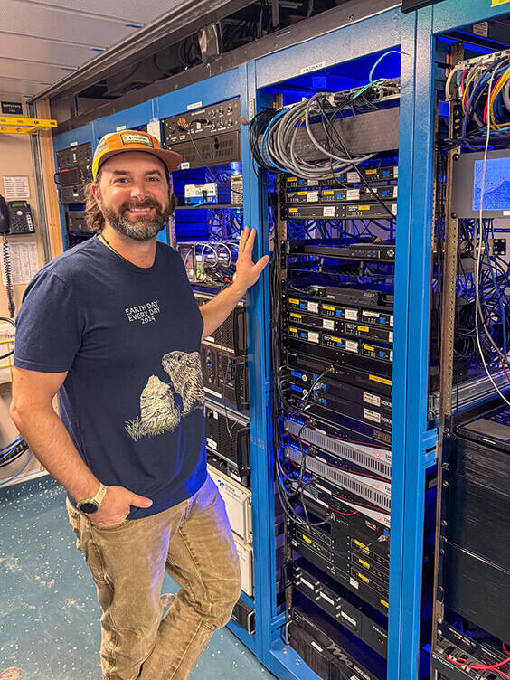 Smiling man in 'Earth Day Every Day 2024' shirt, orange hat, beard, points to glowing blue server racks.
