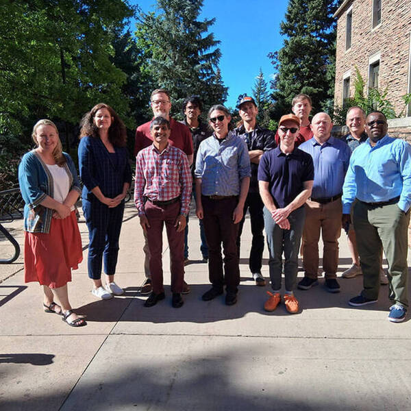 Twelve smiling adults stand on a sunny campus path, backed by a brick building, low stone wall, and trees.