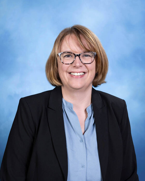 A smiling woman with short blonde hair and tortoiseshell glasses, wearing a black blazer over a blue shirt.
