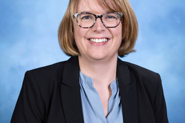 A smiling woman with short blonde hair and tortoiseshell glasses, wearing a black blazer over a blue shirt.