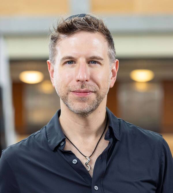 A man with short brown hair and stubble wears a dark shirt and a snake pendant.